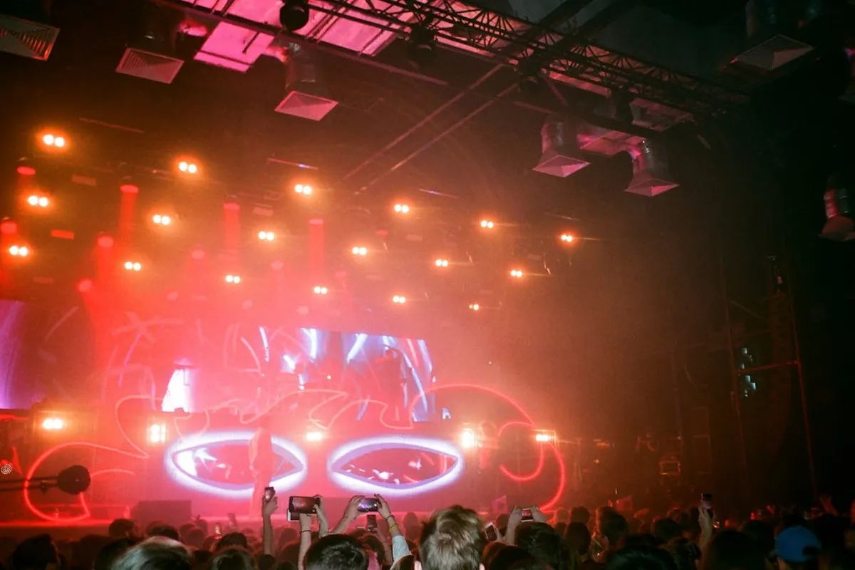 Palco de show ou concerto em ambiente escuro, com público erguendo celulares, iluminado por um telão LED e luzes vermelhas e laranjas dramáticas, representando eventos com tecnologia de iluminação LED.
