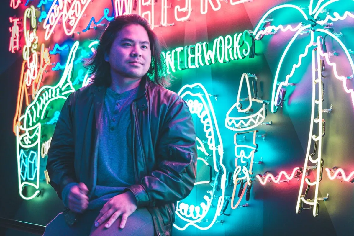 Homem de cabelos longos e jaqueta de couro sentado em frente a um letreiro de LED neon brilhante e colorido, que apresenta desenhos de palmeiras, chapéu mexicano e textos, em um ambiente noturno.
