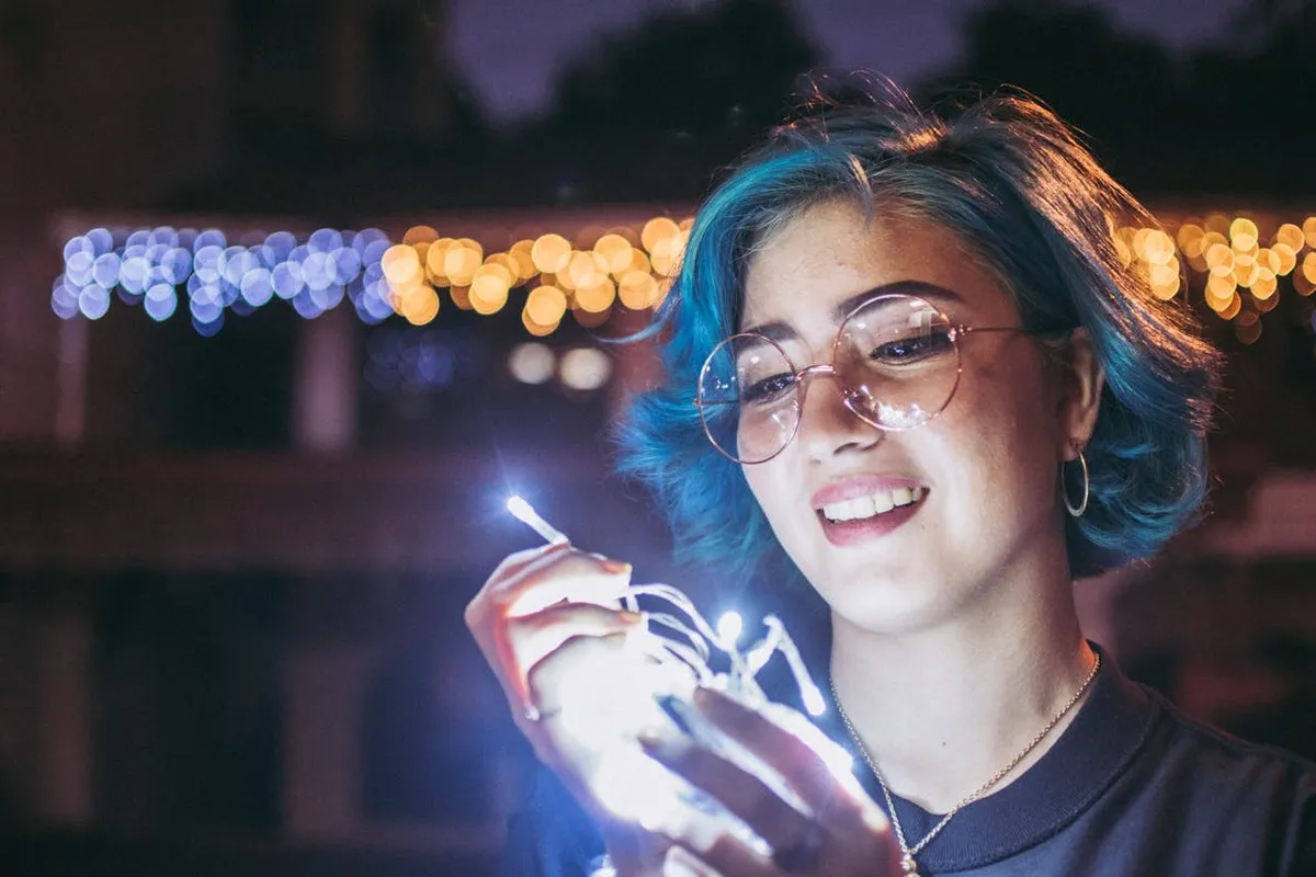 Retrato noturno de close-up de uma jovem sorridente com cabelo azul e óculos de aro redondo, segurando e sendo iluminada por um fio de luzes de LED brancas.