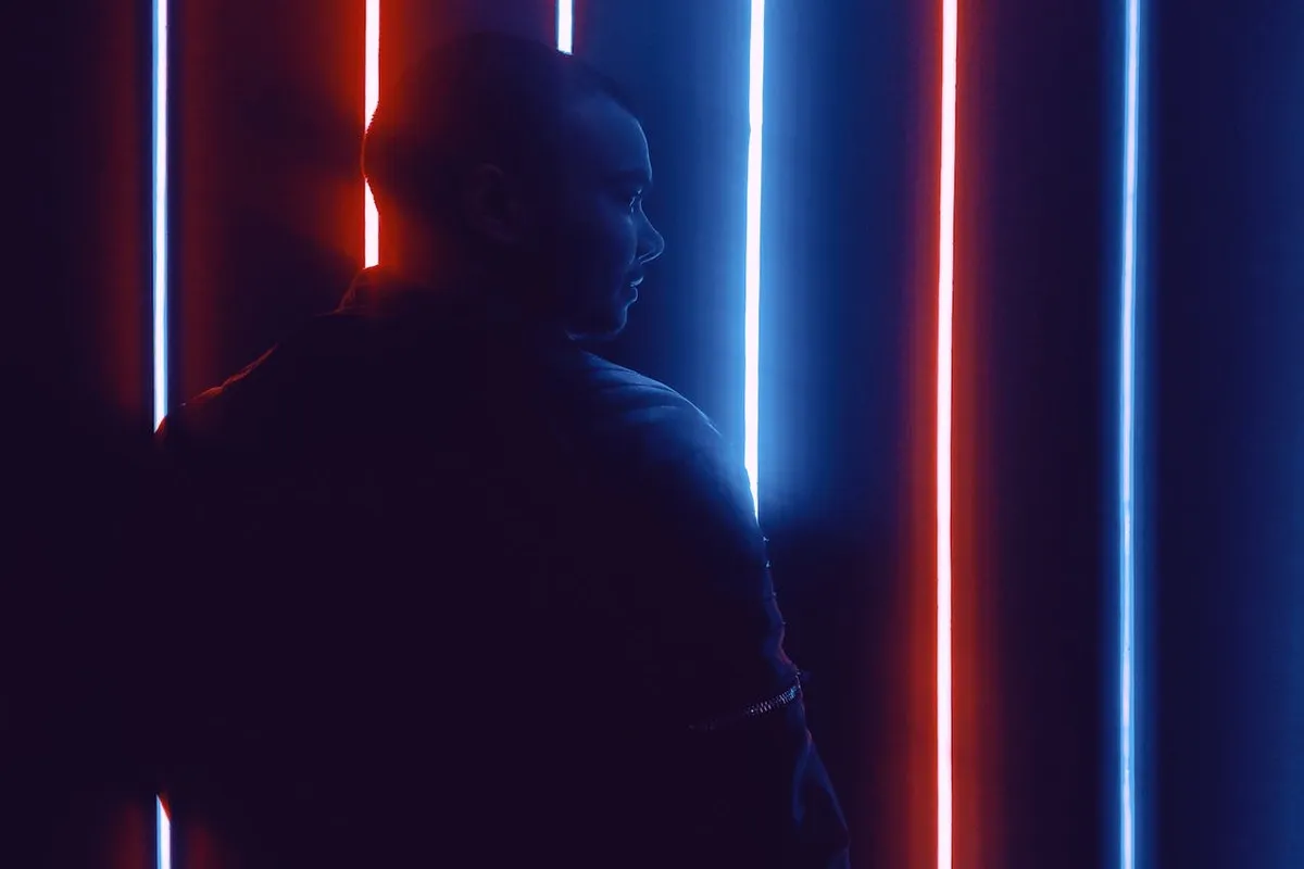 Silhueta de um homem de perfil contra luzes verticais de LED ou neon nas cores azul e vermelho-alaranjado, criando um efeito de contraluz dramático e moderno.