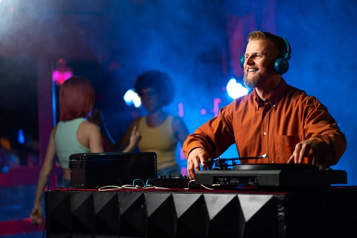 DJ sorridente usando fones de ouvido, operando toca-discos em uma festa com iluminação colorida e pessoas dançando ao fundo.