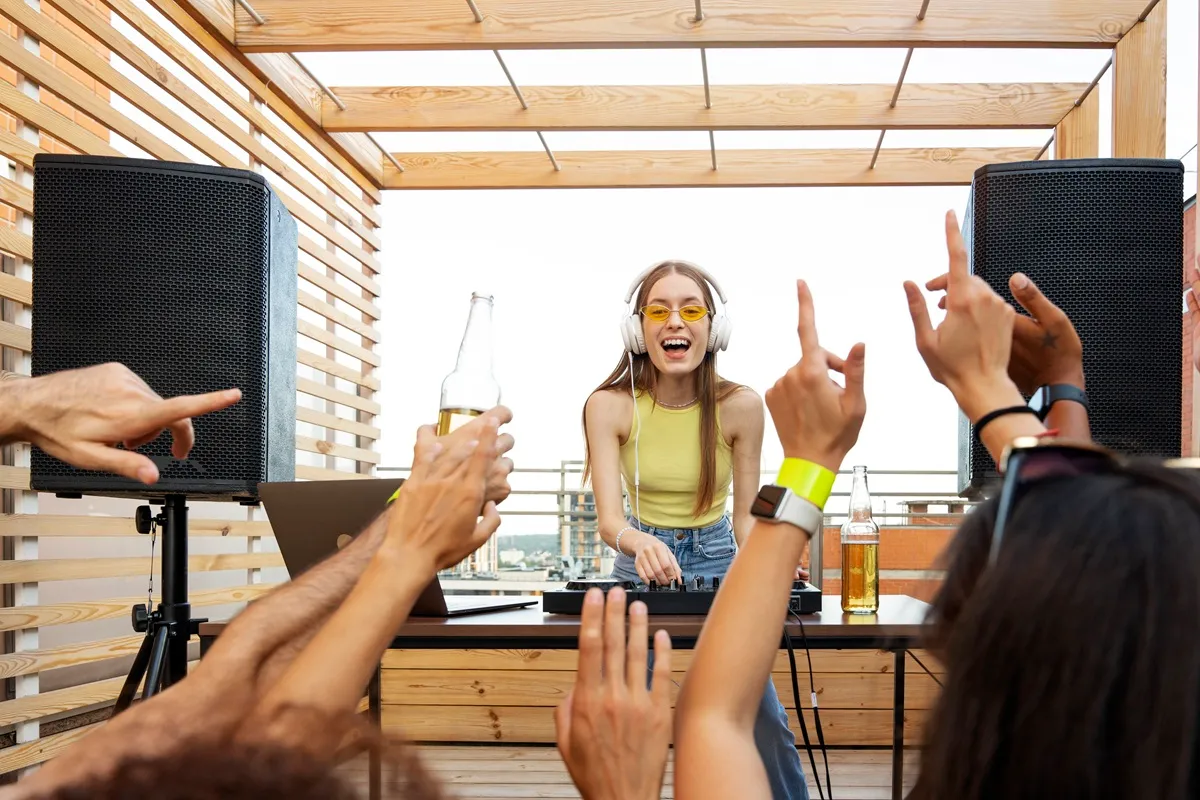 Jovem DJ sorridente usando fones de ouvido e óculos amarelos, tocando música em festa ao ar livre com caixas de som grandes e pessoas dançando e comemorando.