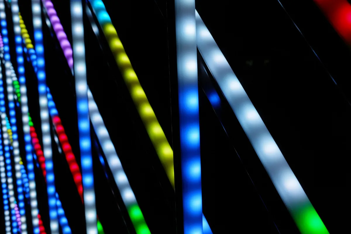 Close-up em ângulo diagonal de barras ou fitas de LED verticais em um fundo escuro, com cores vibrantes como azul, branco, amarelo, vermelho e verde, criando um efeito de luz intensa e moderna.