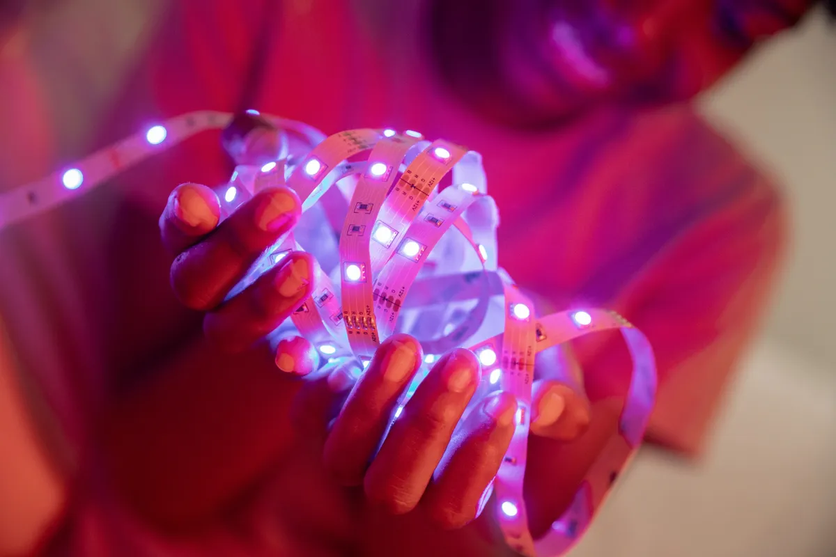 Close-up das mãos de uma pessoa segurando uma tira enrolada de fita de LED (LED strip) que brilha intensamente em luz branca e azul, com reflexos em tons de magenta e rosa no fundo, ideal para iluminação decorativa e DIY.