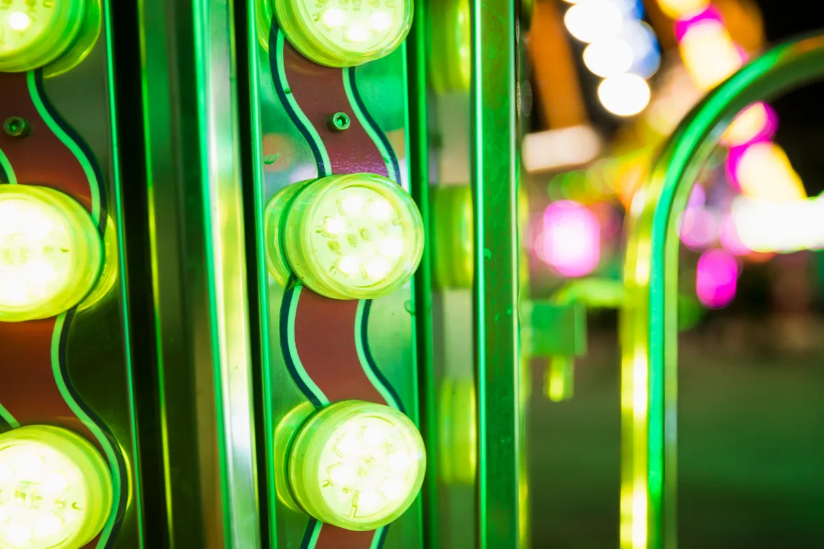 Close-up de lâmpadas de LED circulares de luz amarela/verde-limão intensa, montadas em uma superfície metálica vermelha e verde, provavelmente em um brinquedo de parque de diversões ou carnaval, com luzes bokeh ao fundo.