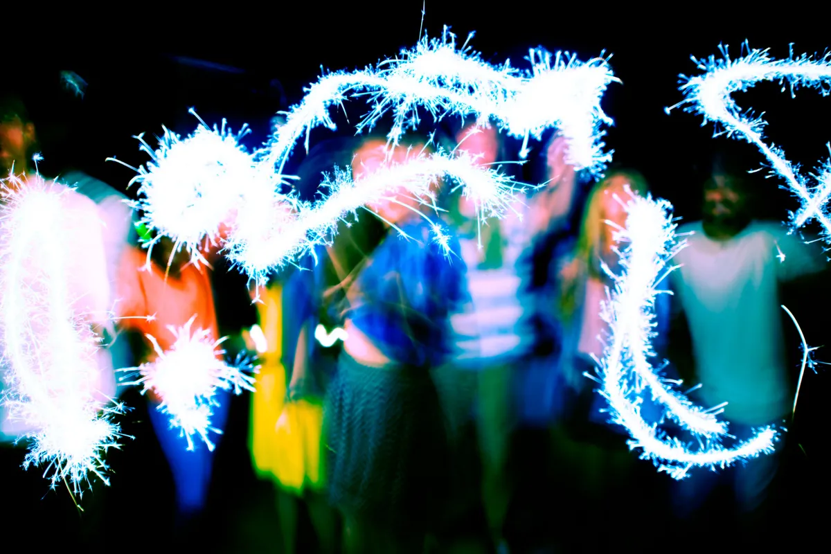 Foto de longa exposição com rastros de luz de sparklers (vela faísca) brancos e brilhantes, desenhados no ar por um grupo de jovens que aparecem desfocados, simbolizando iluminação criativa e celebração noturna.