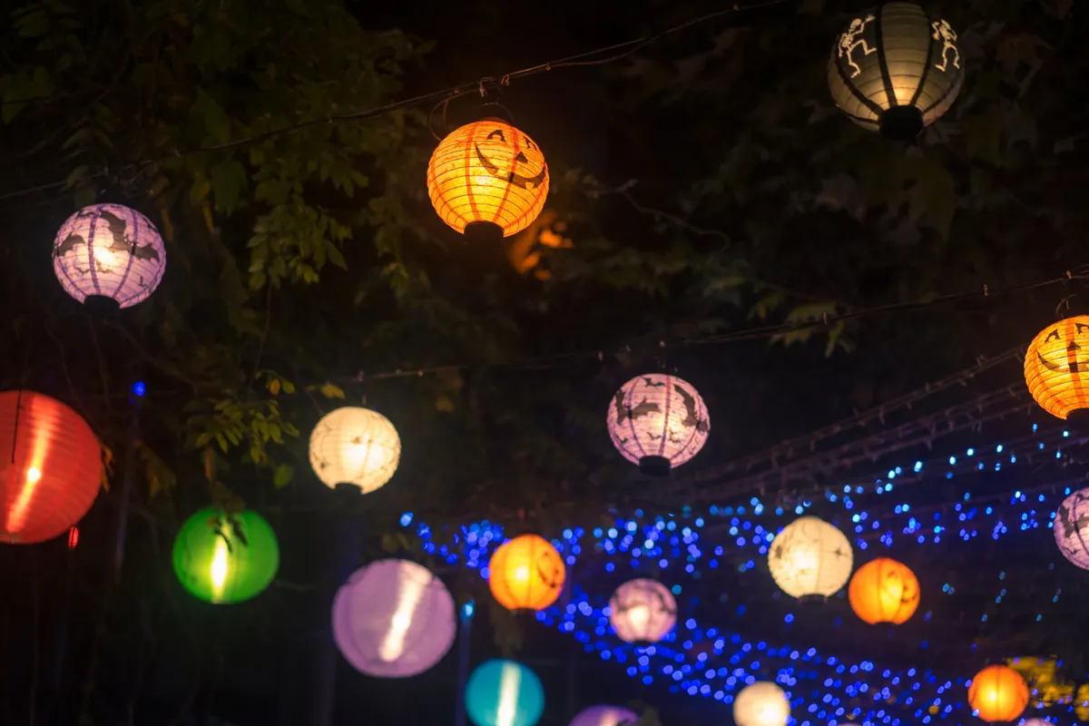 Decoração noturna com uma guirlanda de lanternas de papel redondas, penduradas em fios, em várias cores (laranja, roxo, verde, branco), misturadas com luzes de LED azuis cintilantes, criando uma atmosfera de festa.