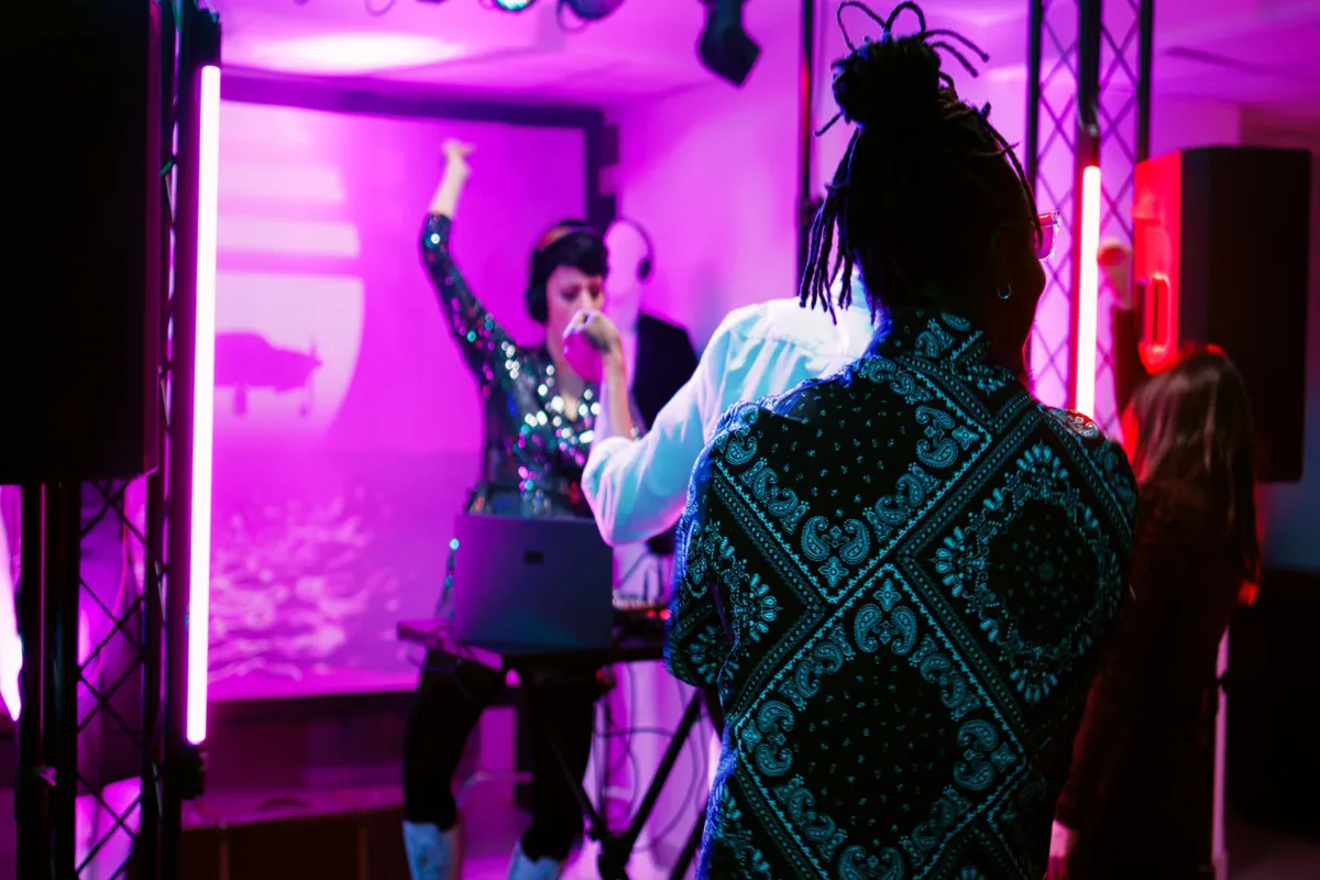 Cena de festa ou show em ambiente interno com forte iluminação rosa e magenta, onde uma DJ feminina está mixando música e é observada por pessoas, destacando o uso de iluminação cênica.