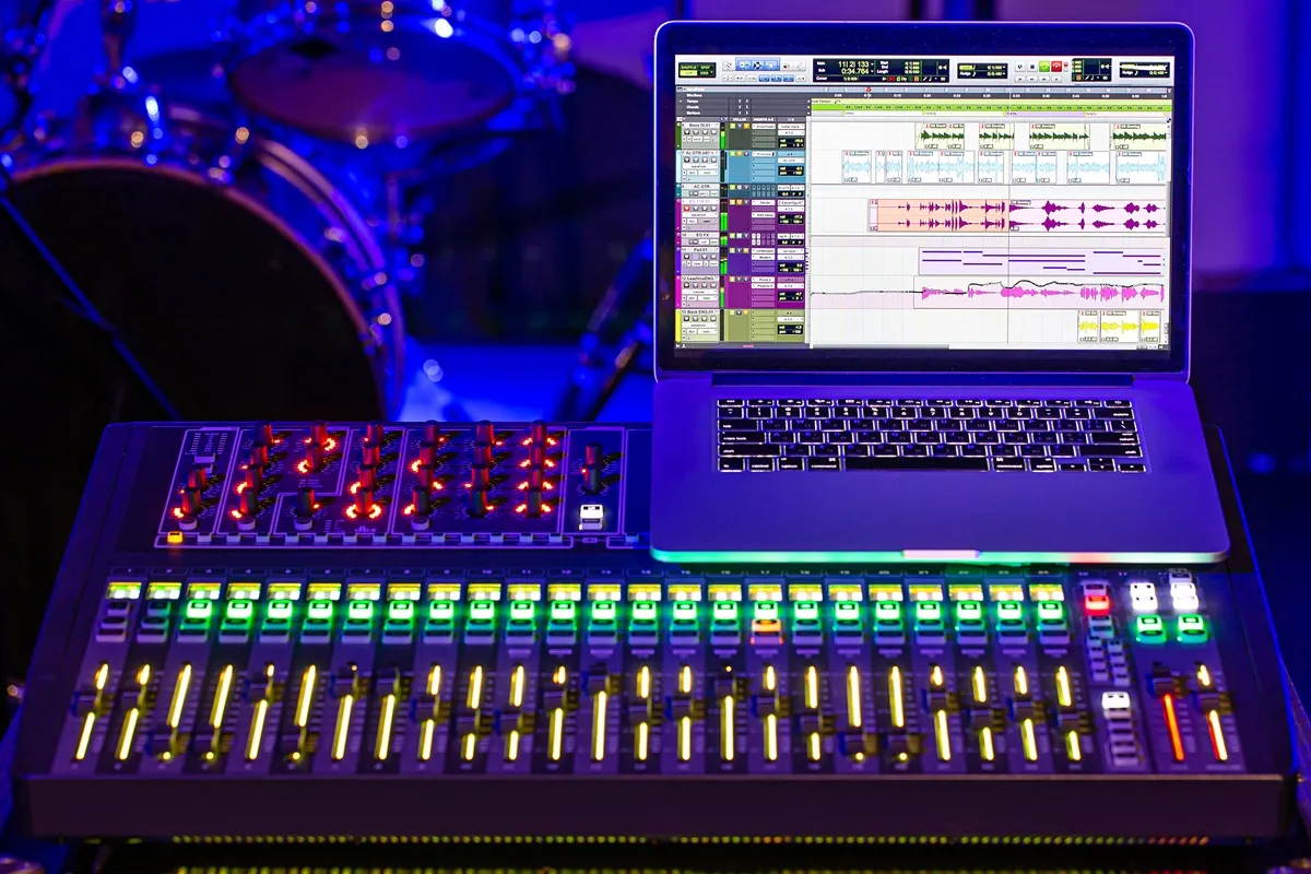 Mesa de som digital iluminada com luzes coloridas, acompanhada de notebook aberto mostrando software de edição de áudio em um estúdio de música.