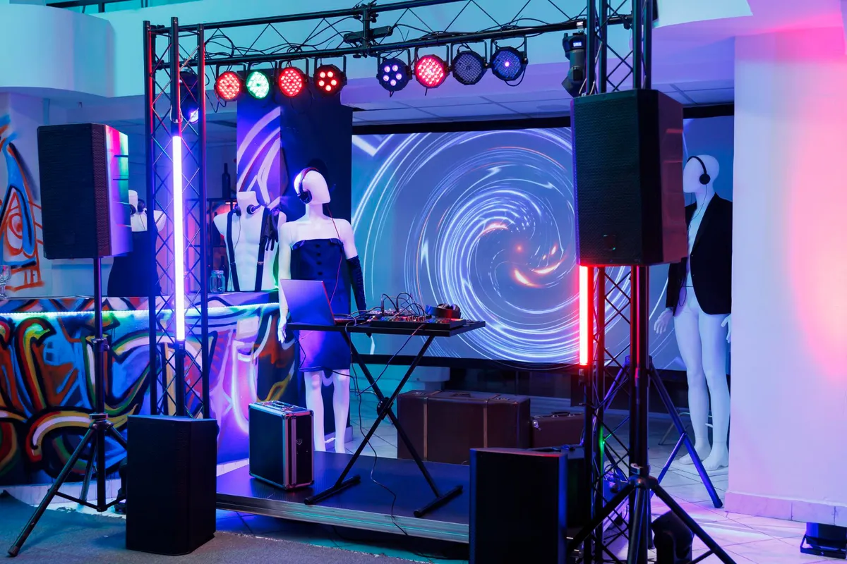 Palco de DJ montado para um evento com manequins, grandes caixas de som, iluminação de LED azul e rosa e um painel de LED ao fundo exibindo um gráfico abstrato em espiral.