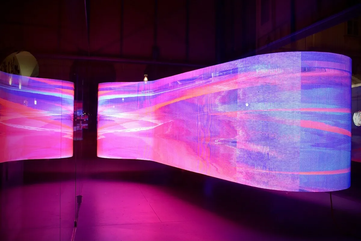 Telão de LED curvo e ondulado exibindo um gráfico abstrato em movimento nas cores magenta, rosa, roxo e azul, instalado em um ambiente escuro, simulando uma instalação artística ou display de evento.