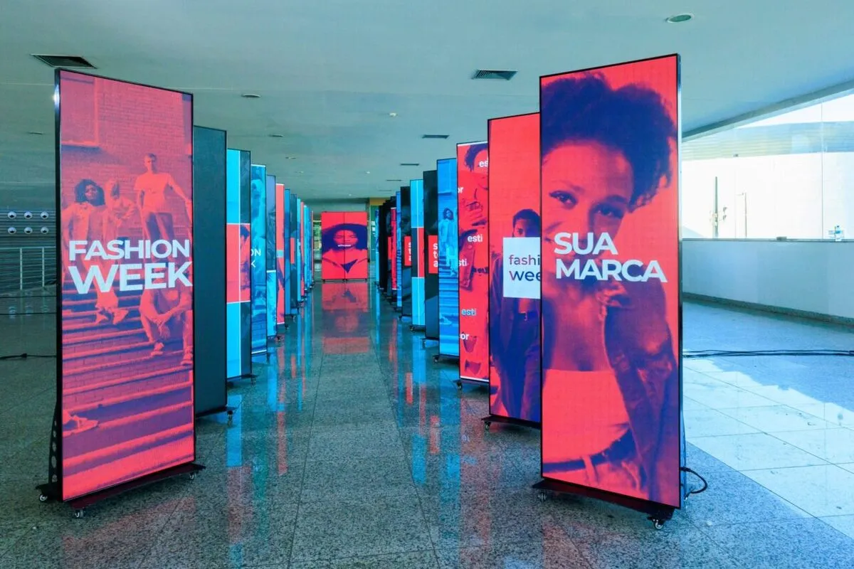 Corredor iluminado com uma série de totens de LED verticais em ângulo de perspectiva, exibindo imagens de moda em tons de vermelho e azul, com textos como "FASHION WEEK" e "SUA MARCA", ideal para sinalização de eventos.