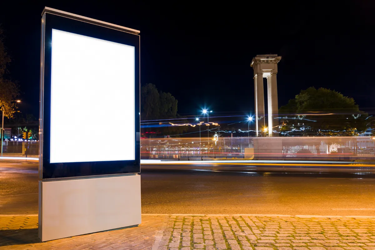 Totem digital de LED vertical com tela branca vazia (mockup) em primeiro plano, posicionado na calçada de uma rua urbana à noite, com rastros de luz de veículos em longa exposição.