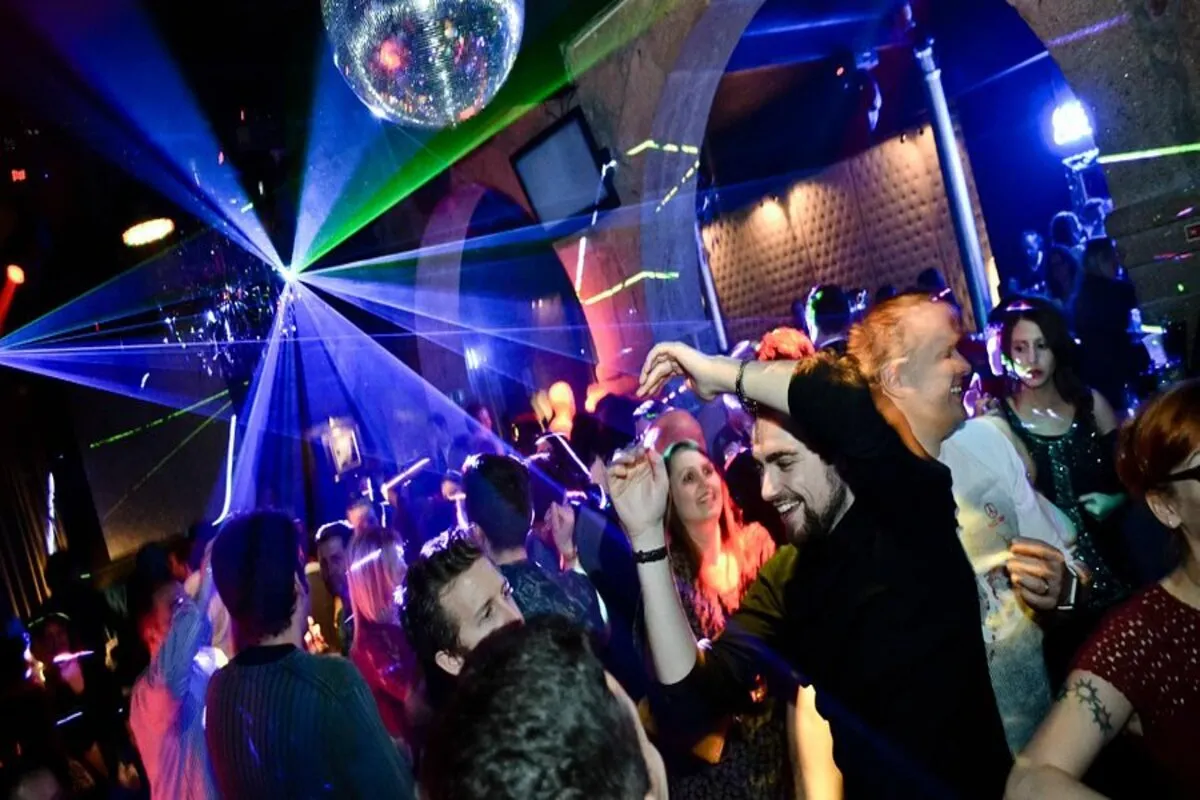  Pista de dança de boate com pessoas em movimento, iluminada por um globo espelhado (disco ball), lasers azuis e verdes, e luzes vermelhas, criando uma atmosfera noturna e energética.