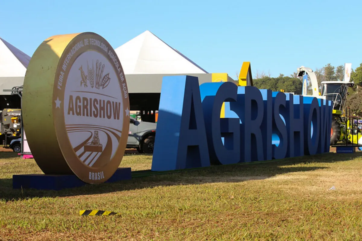 Letreiro da Agrishow em destaque na entrada da feira, com o logotipo dourado e letras azuis grandes sobre a grama, em um dia ensolarado de evento agrícola.