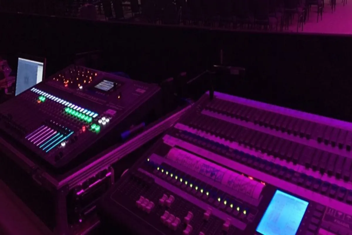 Close-up de duas mesas de som digital em um ambiente de palco escuro, iluminadas por luzes roxas e azuis, com vários faders e botões.