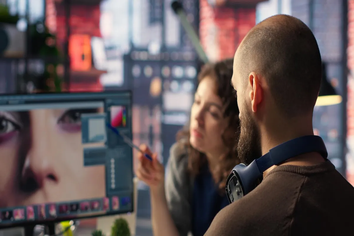 Close-up de dois editores de vídeo ou designers gráficos em uma sala escura, com um homem de costas e uma mulher apontando para detalhes em um monitor que exibe o close-up de um olho.