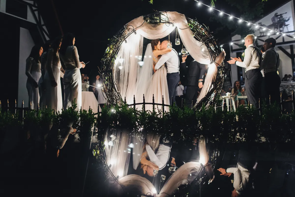 Casamento noturno ao ar livre com noivos se beijando sob um arco de galhos e luzes, perfeito para eventos românticos.