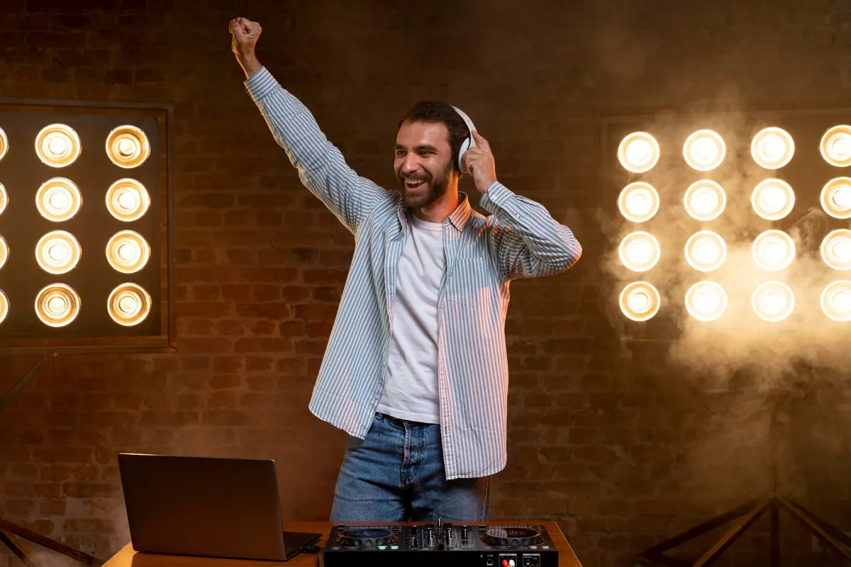 Um DJ feliz e sorridente, com fones de ouvido brancos, levanta um braço no ar em uma celebração enquanto mixa música em um console de DJ com um laptop. O fundo é iluminado por vários holofotes em LED que criam uma atmosfera energética e esfumaçada.