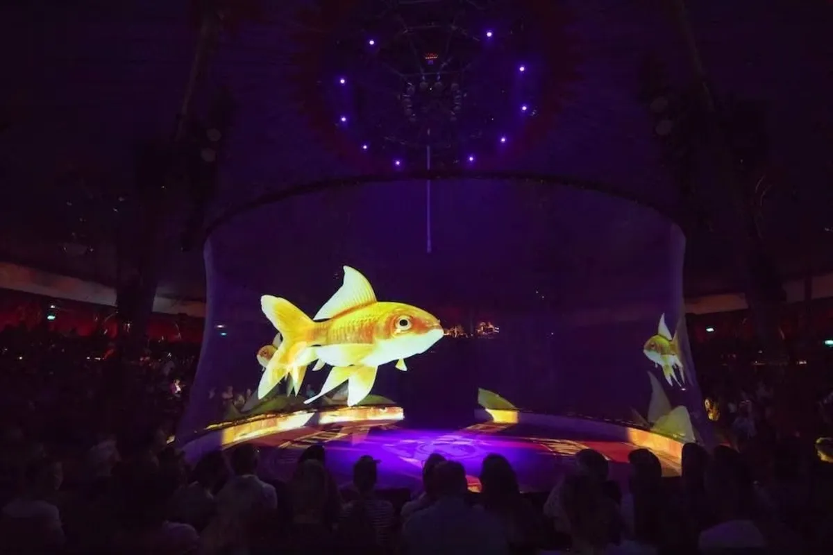 Projeção holográfica 3D de um grande peixe-dourado flutuando em um aquário virtual no centro de um picadeiro de circo ou palco com plateia, usando iluminação azul e roxa.