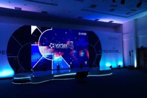 Palco de evento corporativo com grande telão de LED em formato curvo e projetor 4K, exibindo o logo "Vortex 15 Anos" em um design futurista de luz azul.