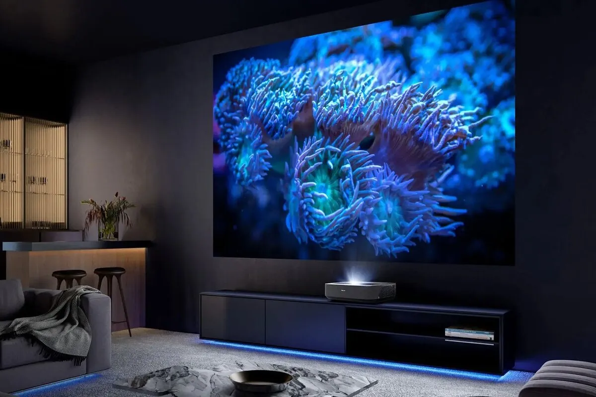 Projetor de ultracurta distância exibindo uma imagem 4K vibrante e luminosa de anêmonas marinhas azuis em uma tela gigante em uma sala de estar moderna e escura.