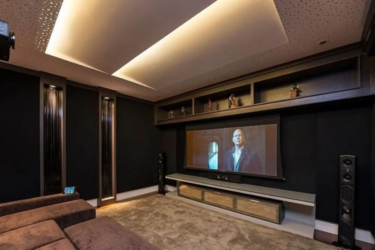 Sala de cinema residencial luxuosa com tela de projeção exibindo um filme, alto-falantes de torre para som ambiente, e painéis acústicos pretos.