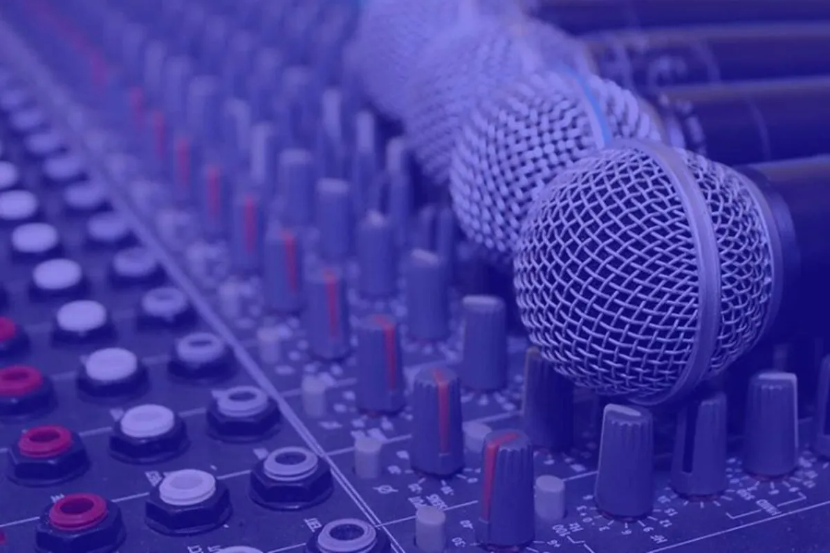 Vários microfones posicionados sobre uma mesa de som estéreo profissional, com botões e controles de áudio em destaque sob iluminação azul.