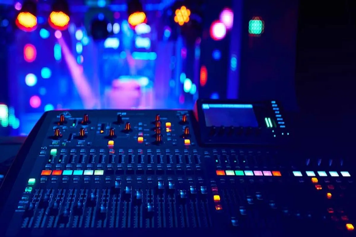 Mesa de som estéreo profissional iluminada por luzes coloridas de palco, com botões e controles acesos em um ambiente de show ou estúdio musical.