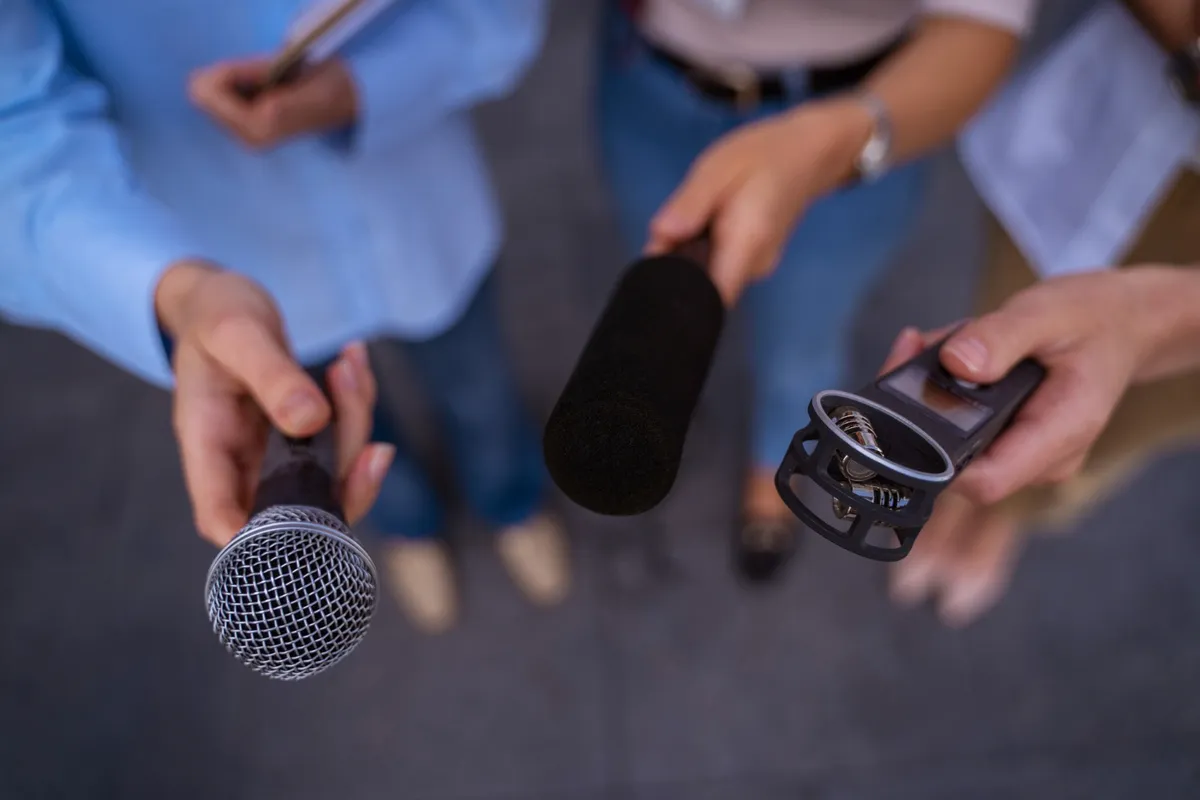 Vista de cima de repórteres segurando três tipos de microfone: microfone dinâmico de mão, microfone shotgun com espuma e um gravador digital portátil para entrevista.