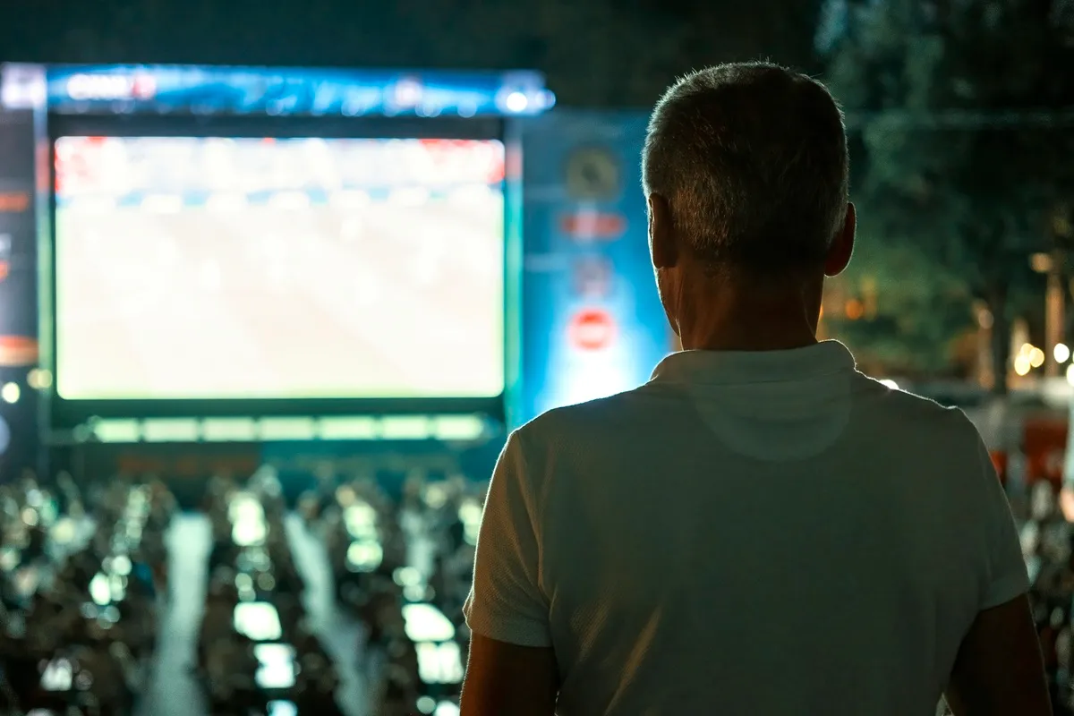 Homem visto de costas observando um grande telo de LED exibindo jogo em evento noturno ao ar livre.