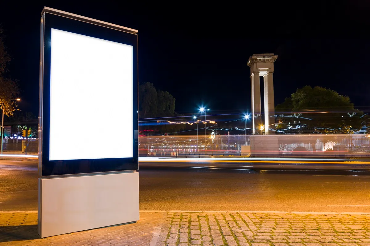 Painel de LED vazio iluminado em uma avenida ? noite, com luzes de carros em movimento e monumento ao fundo.