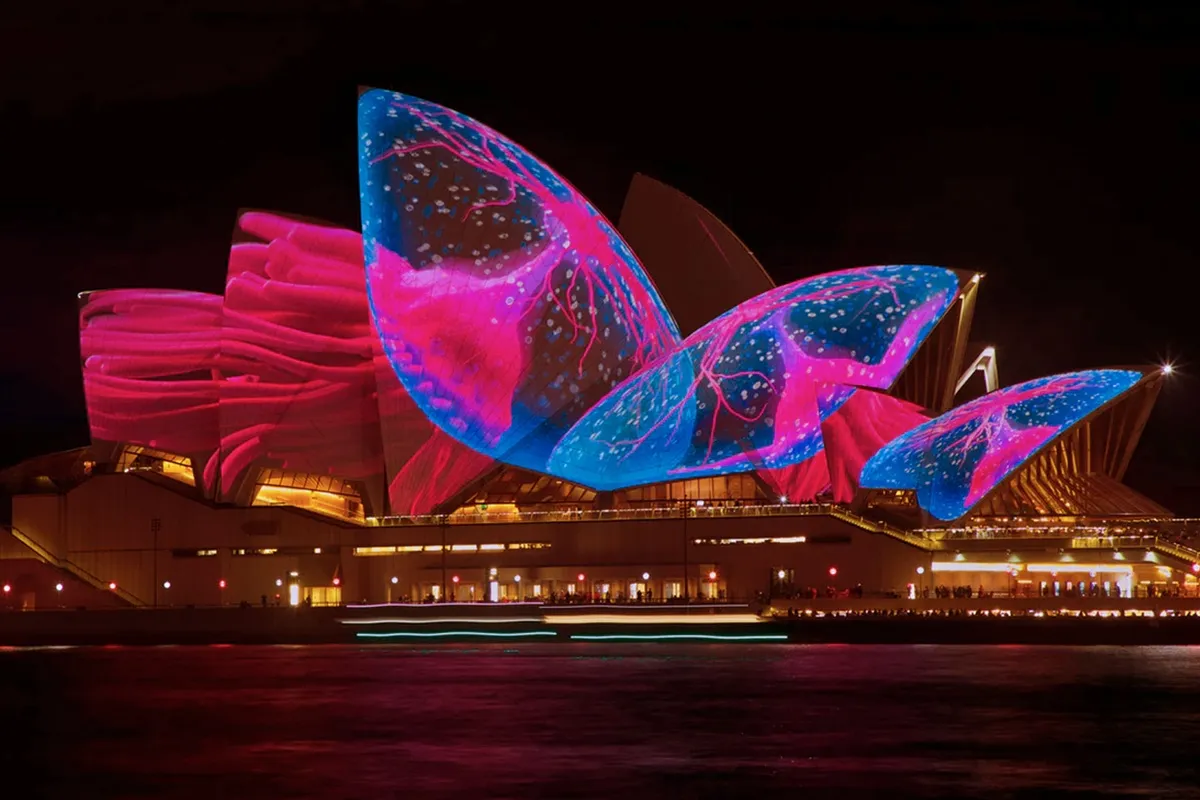 Projeção mapeada colorida ilumina a Ópera de Sydney à noite, com imagens em tons de azul e rosa sobre suas conchas arquitetônicas.