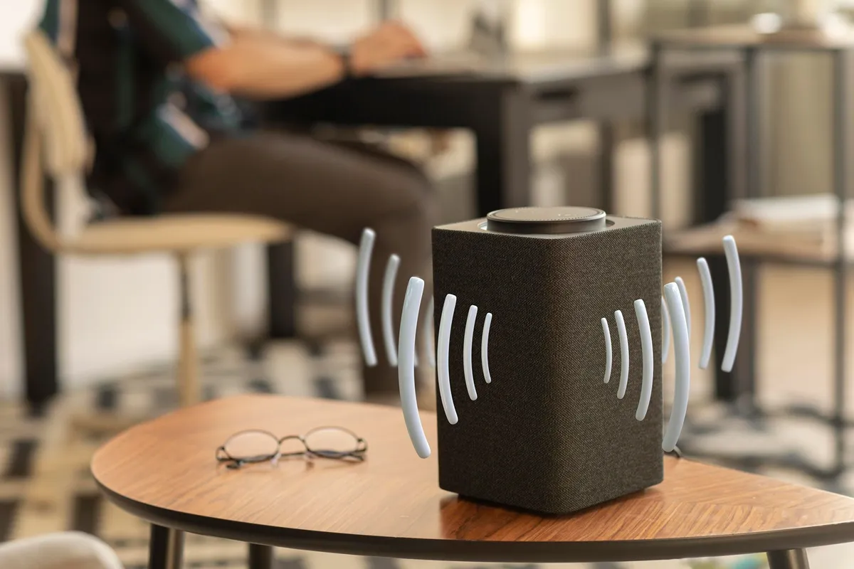 Caixa de som Bluetooth preta emitindo ondas sonoras sobre uma mesa de madeira, com uma pessoa desfocada ao fundo em um ambiente doméstico.