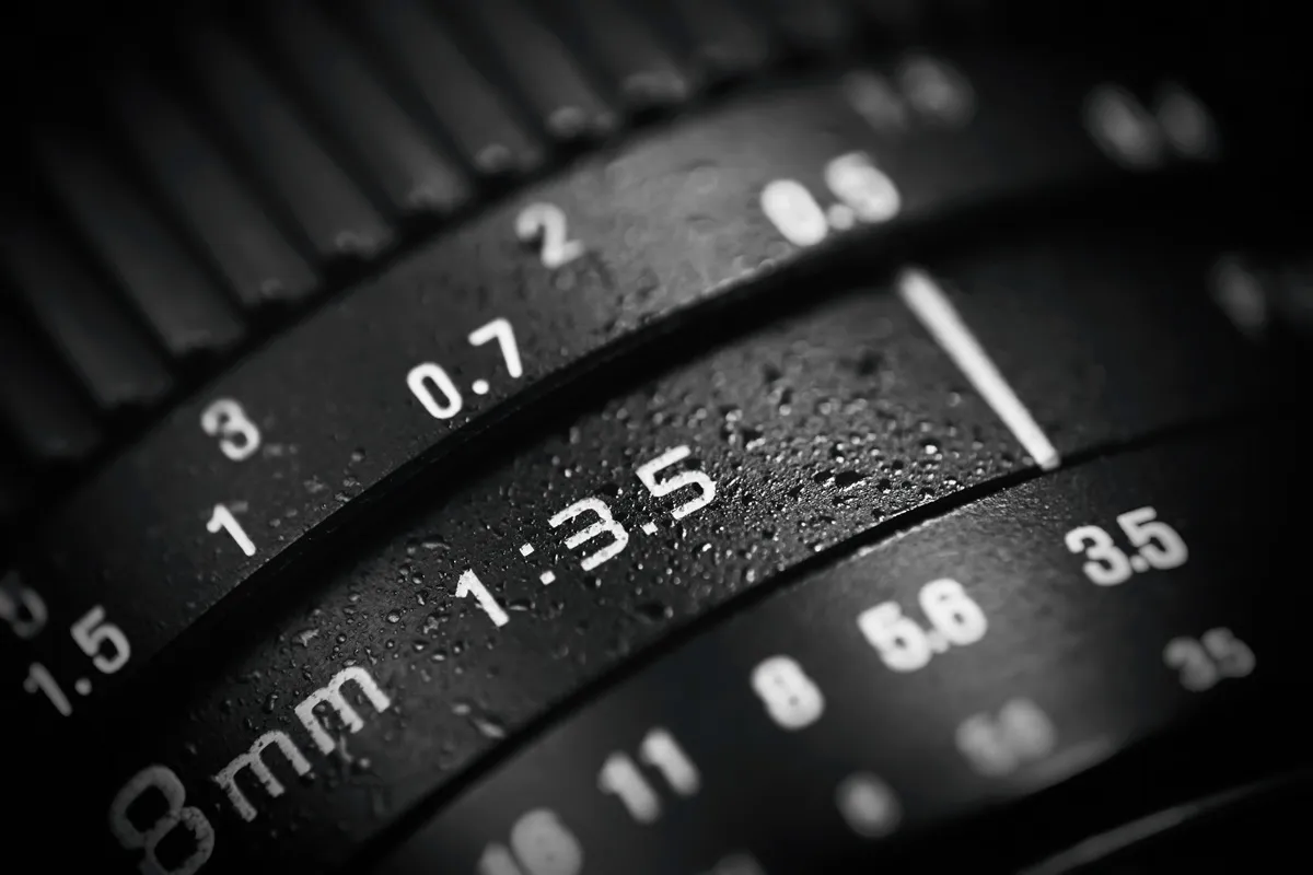 Close-up de lente de câmera fotográfica com marcações de abertura f/1.35 e escala de foco em fundo escuro