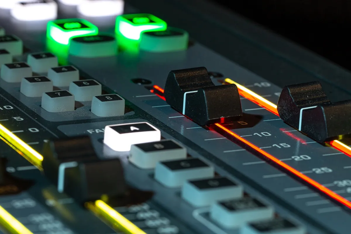 Close-up de uma mesa de som com sliders, botões iluminados e indicadores de nível em um estúdio de áudio.