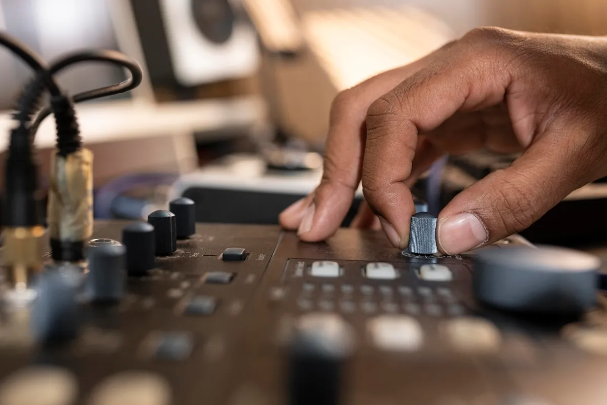 Mão ajustando um knob em uma mesa de som, cercada por botões e cabos em um estúdio de áudio.