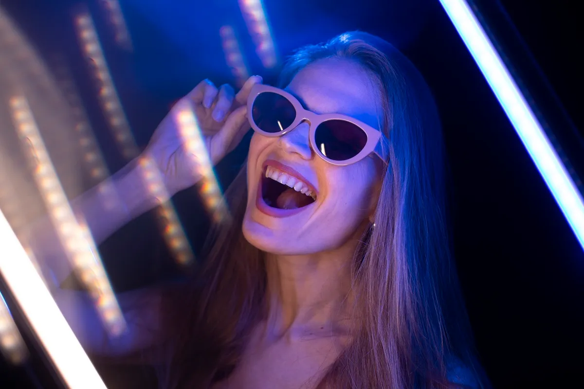 Mulher sorrindo usando óculos estilizados, iluminada por luz UV azul e rosa em um cenário com efeitos de luz neon.