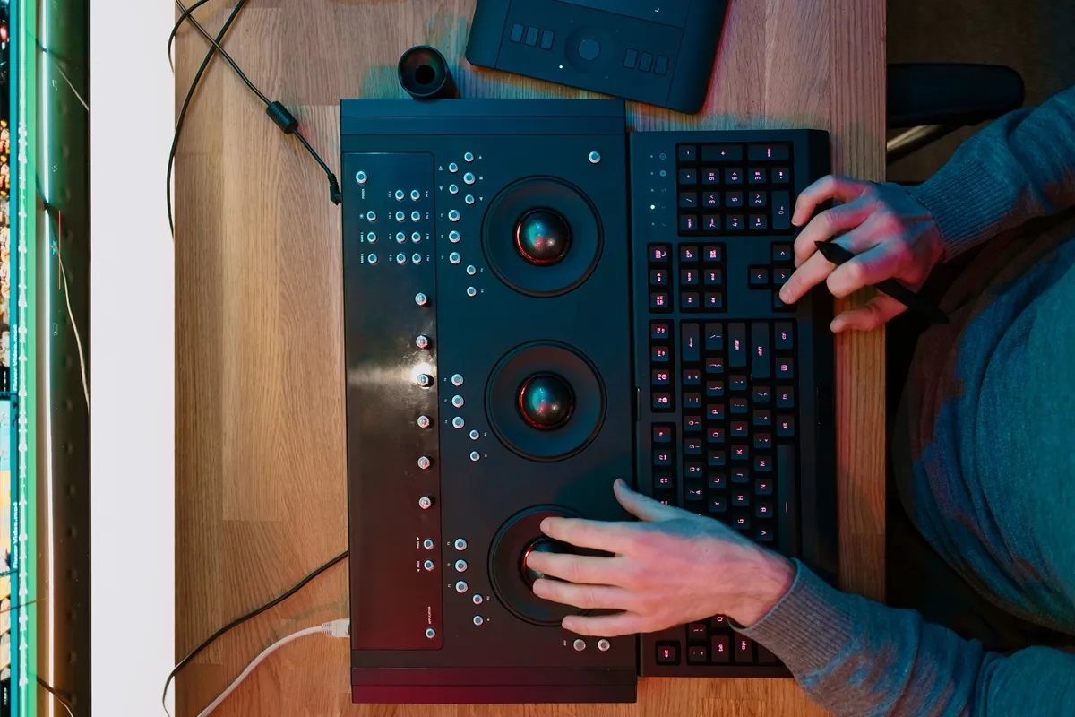 Pessoa operando um painel de correção de cor com esferas e botões ao lado de um teclado iluminado em um ambiente de edição profissional.