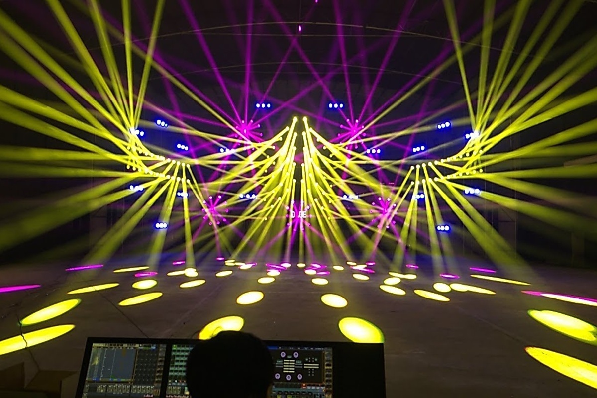Imagem de um show de iluminação profissional com moving heads emitindo feixes de luz amarelos e roxos, formando padrões simétricos sobre um palco amplo.