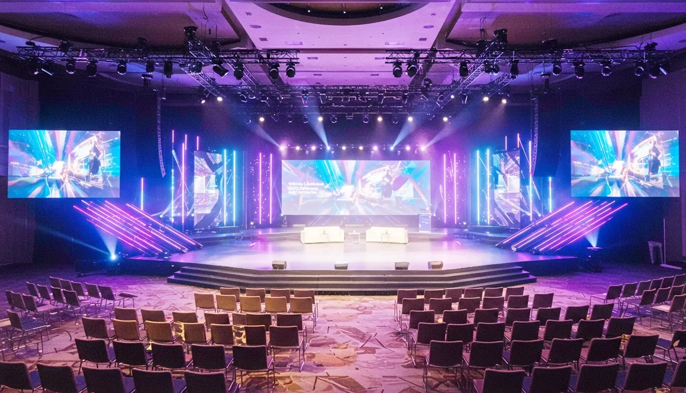 Palco amplo de evento com telões gigantes, luzes LED coloridas e cadeiras organizadas na plateia em ambiente corporativo.
