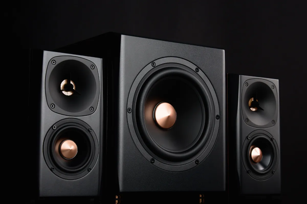 Sistema de som com subwoofer central e duas caixas de som posicionadas ao lado em fundo escuro.