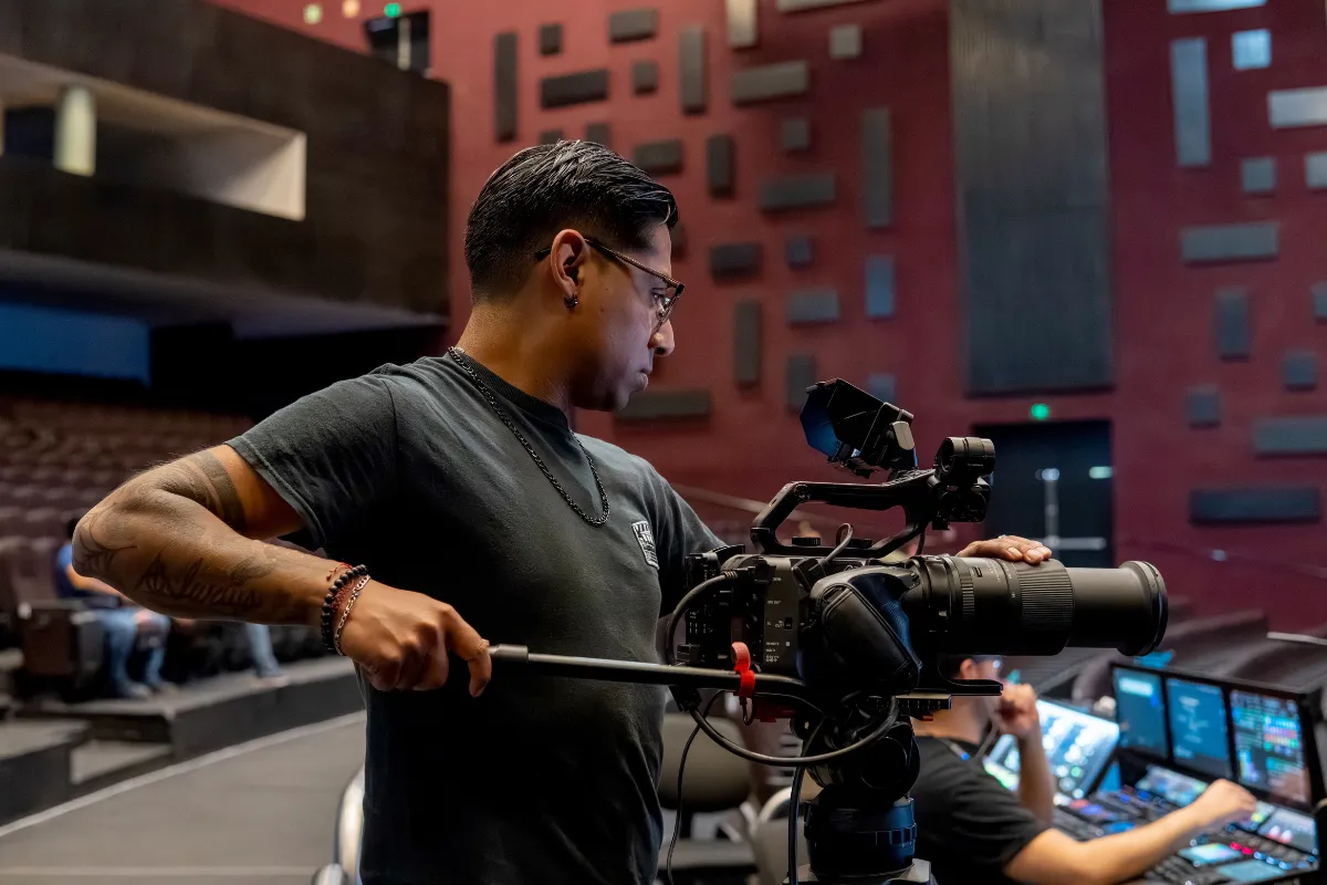 Profissional operando câmera de filmagem em auditório com equipamentos de controle ao fundo