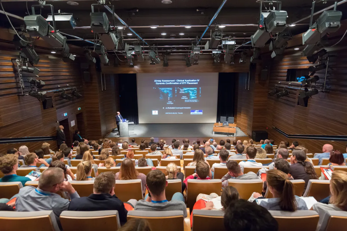Grande auditório moderno com palco, telão e público assistindo a uma apresentação durante um evento corporativo.