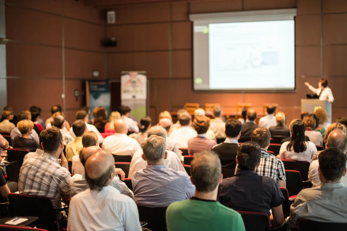 Plateia sentada em auditório assistindo a uma apresentação em tela durante um congresso empresarial.