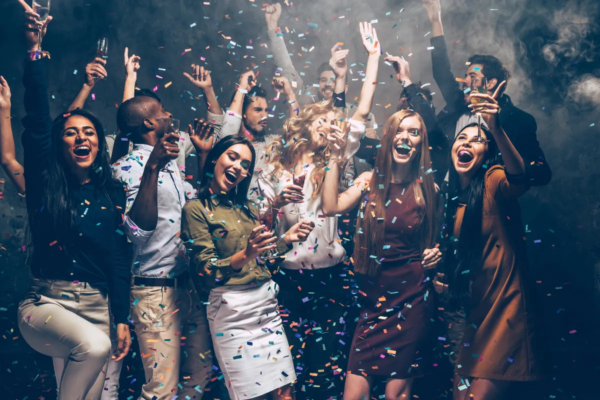 Grupo de pessoas comemorando em uma festa enquanto confetes caem e alguns seguram taças de bebida.