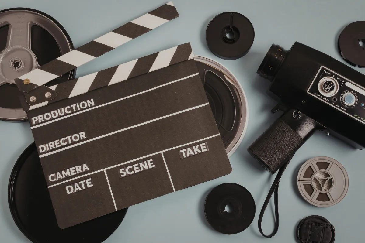 Claquete de cinema sobre mesa ao lado de câmera de filmagem e rolos de filme utilizados em produção audiovisual.