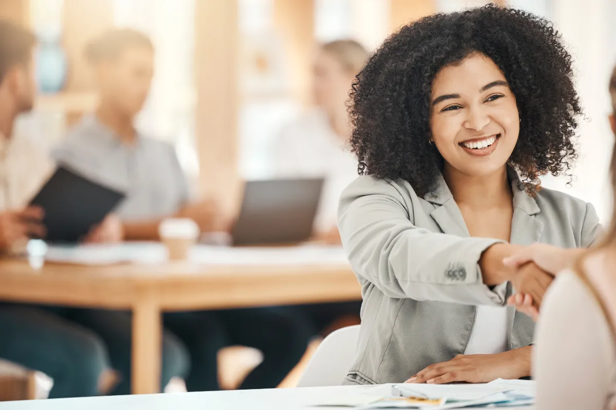 Mulher sorrindo enquanto aperta a mão de outra pessoa em ambiente corporativo com equipe ao fundo