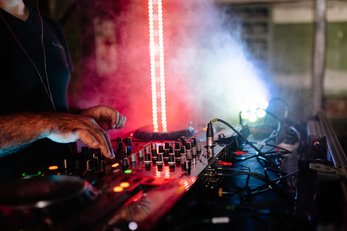 DJ ajustando os controles de uma mesa de som com luzes coloridas e fumaça durante evento noturno.