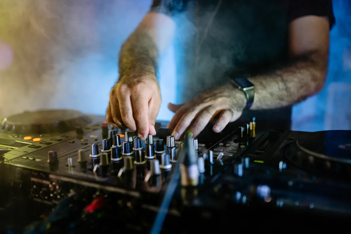 Pessoa ajustando os controles de volume em uma mesa de som profissional durante evento com iluminação e fumaça ao fundo.